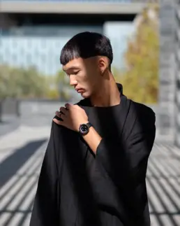 A person wearing a black outfit and a watch, posing outdoors with sunlight casting shadows around them.
