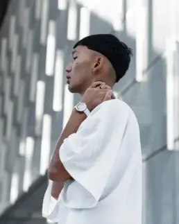 A person in a white shirt with a short, straight haircut and an earring stands sideways, wearing a white wristwatch and holding their neck, against a modern, patterned wall.