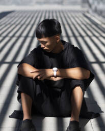 A person sits on a sunlit ground, wearing a black outfit, arms crossed, head down. Shadowed lines create a dramatic pattern, conveying a contemplative mood.