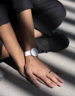 A close-up of hands resting on a gray surface, featuring a modern white wristwatch on one wrist, with soft shadows cast.