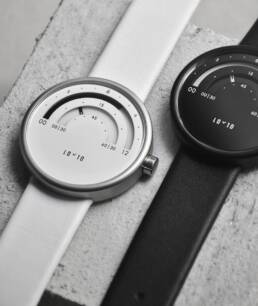 Two minimalist watches on a textured surface; one has a white face with a white strap, the other a black face with a black strap, both showing 10:10.