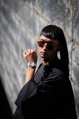 A person dressed in black leans against a textured wall with a minimalist watch visible on their wrist.