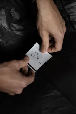 Close-up of two hands holding a small white card with minimalist black text reading ‘LIK–’ against a dark background.