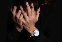 Person covering their face with both hands in a gesture of distress or stress, wearing dark clothing, with a white wristwatch visible on their wrist against a dark background.